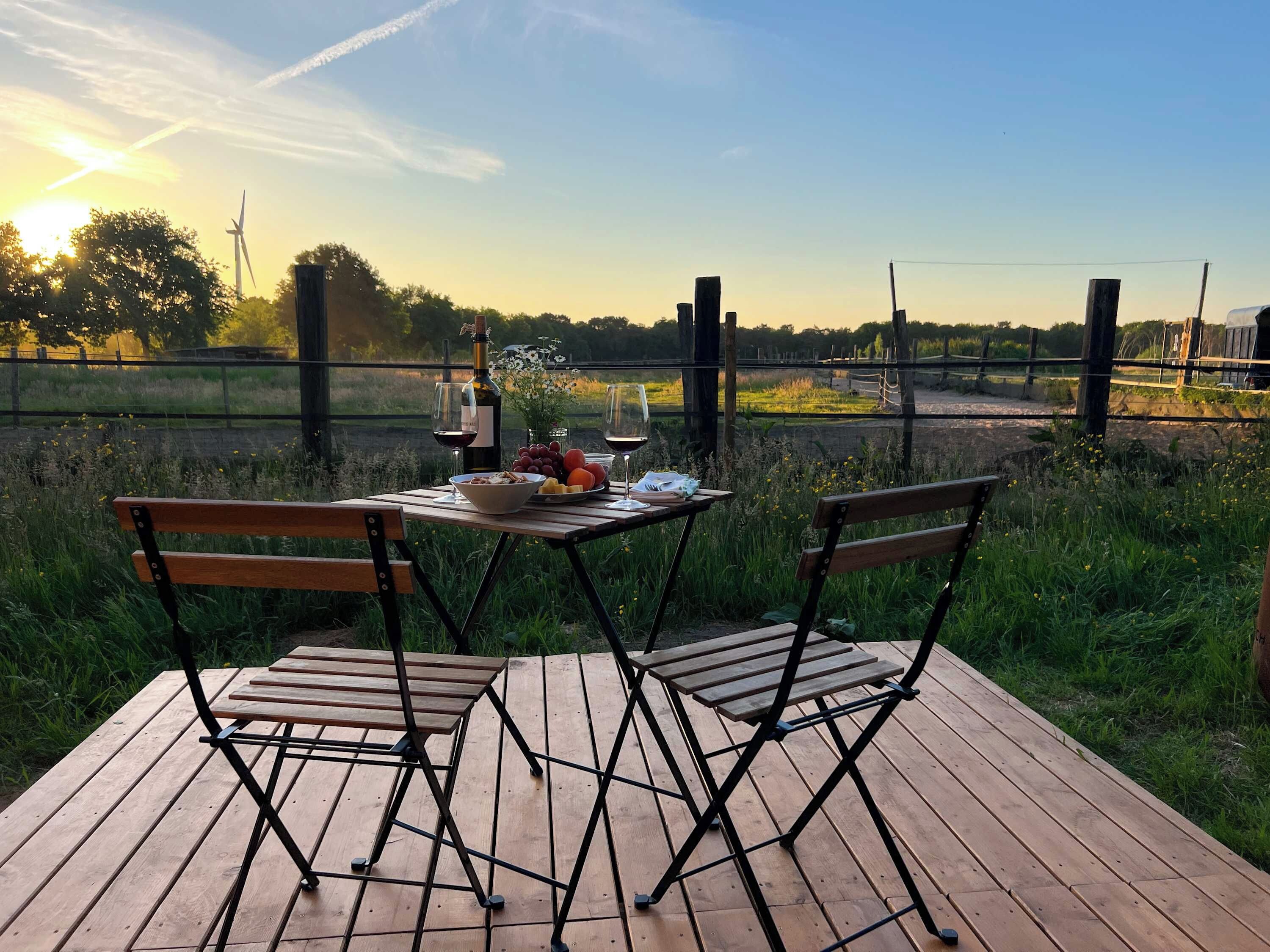 A wooden deck with two chairs and a table set for a meal, overlooking a grassy field and a wind turbine.
