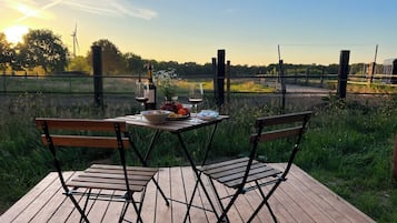 A wooden deck with two chairs and a table set for a meal, overlooking a grassy field and a wind turbine.