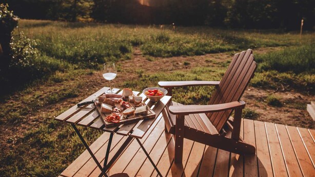 Una terraza de madera con una mesa dispuesta para una comida, una copa de vino blanco y un bol de tomates cherry.