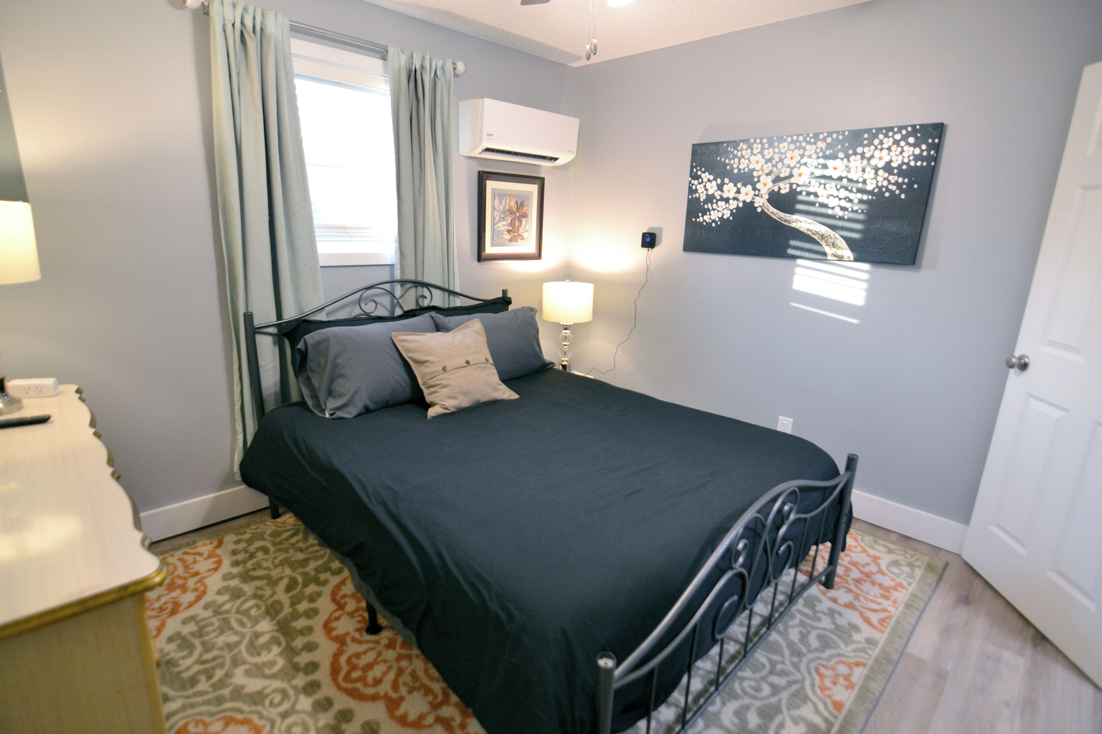 A bedroom with a bed, a bedside table, a lamp, a wall-mounted air conditioner, a framed picture, and a door.