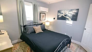 A bedroom with a bed, a bedside table, a lamp, a wall-mounted air conditioner, a framed picture, and a door.
