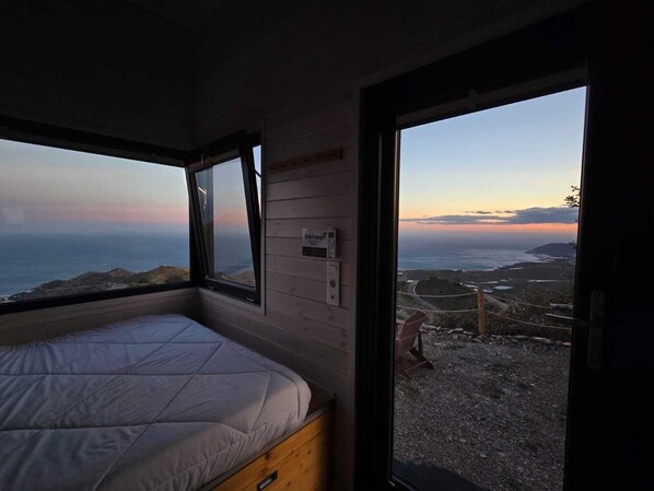 A bedroom with a bed, a window, and a view of the ocean at sunset.