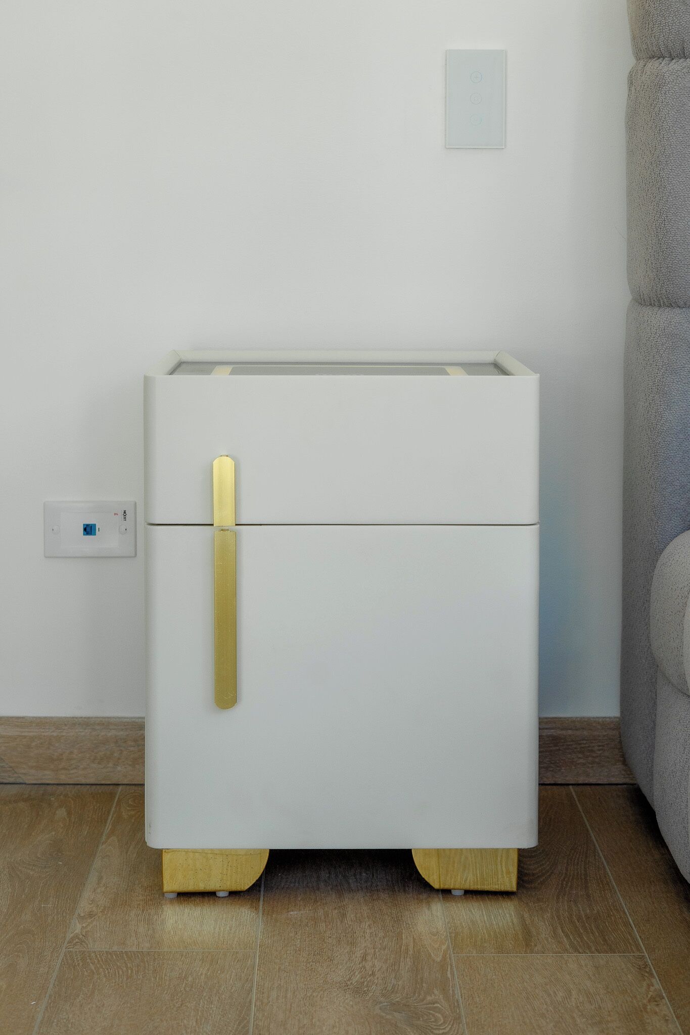 Una mesita de noche blanca con un pomo dorado, un enchufe y un termostato en la pared.