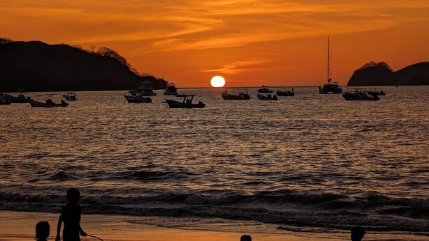 Un coucher de soleil sur une étendue d’eau, avec des bateaux et la silhouette d’une personne sur la rive.