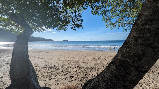 A beach with visible footprints, a clear sky, and boats in the water.