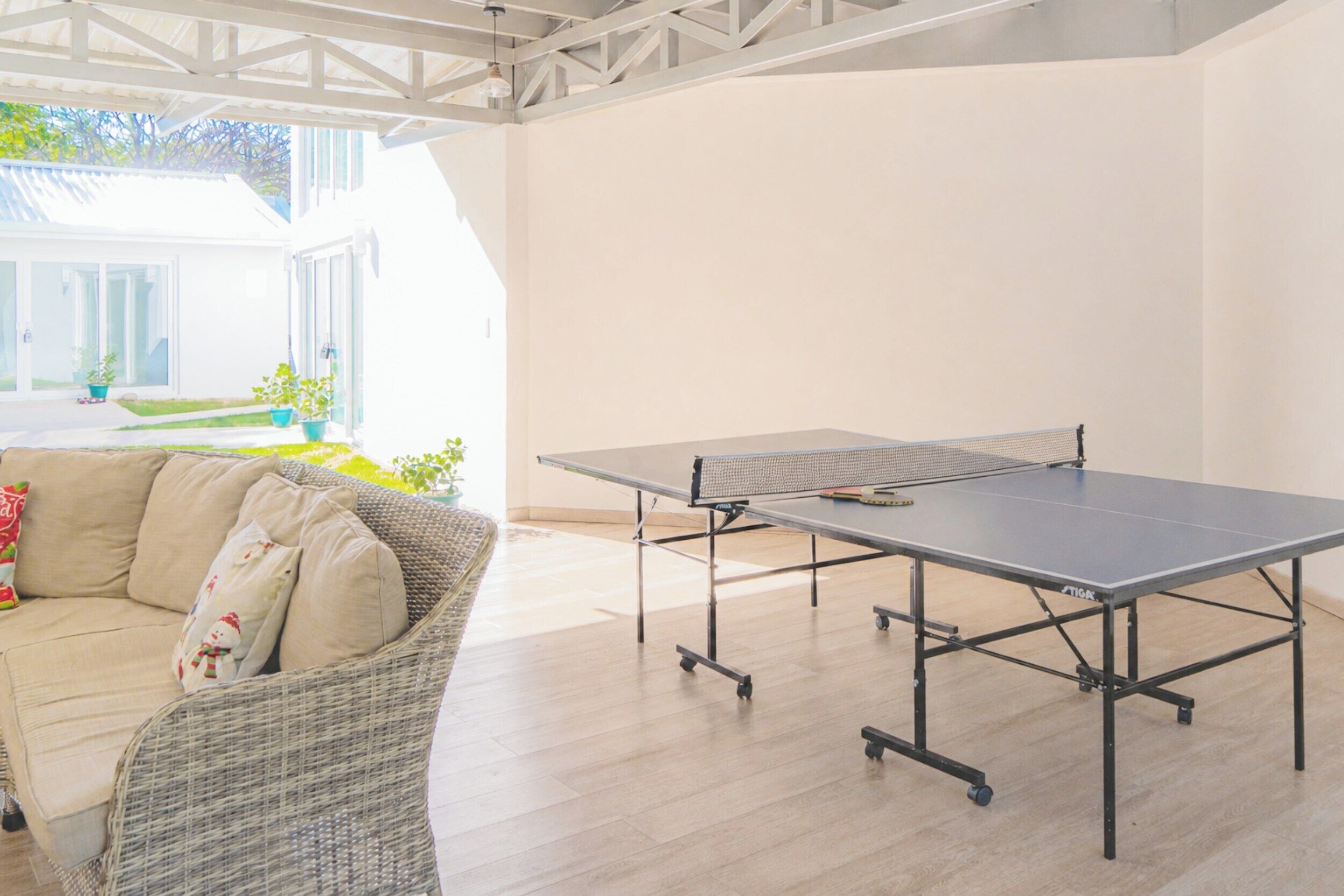 A room with a ping pong table, a wicker sofa, and a view of a garden.