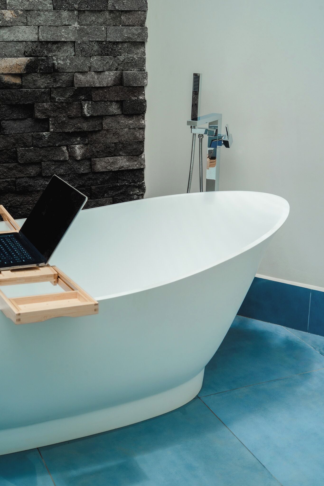 Une salle de bain moderne avec une baignoire sur pieds, un ordinateur portable posé sur un plateau en bois et un mur en pierre.