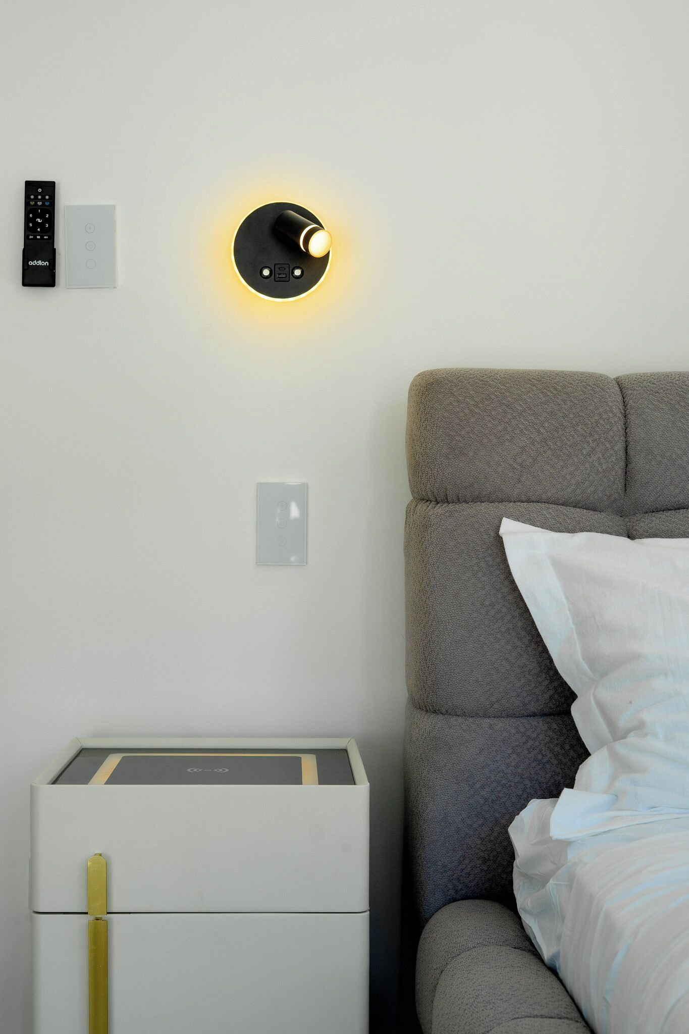 A modern bedroom with a grey upholstered headboard, a bedside table with a lamp, and a wall-mounted control panel.