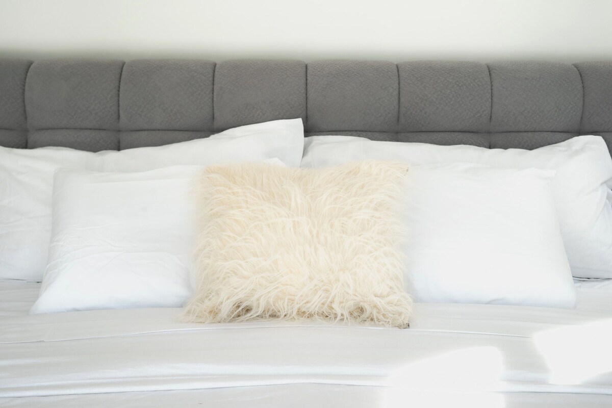 Een bed met een grijs beklede hoofdbord, wit beddengoed en een pluizig beige kussen.