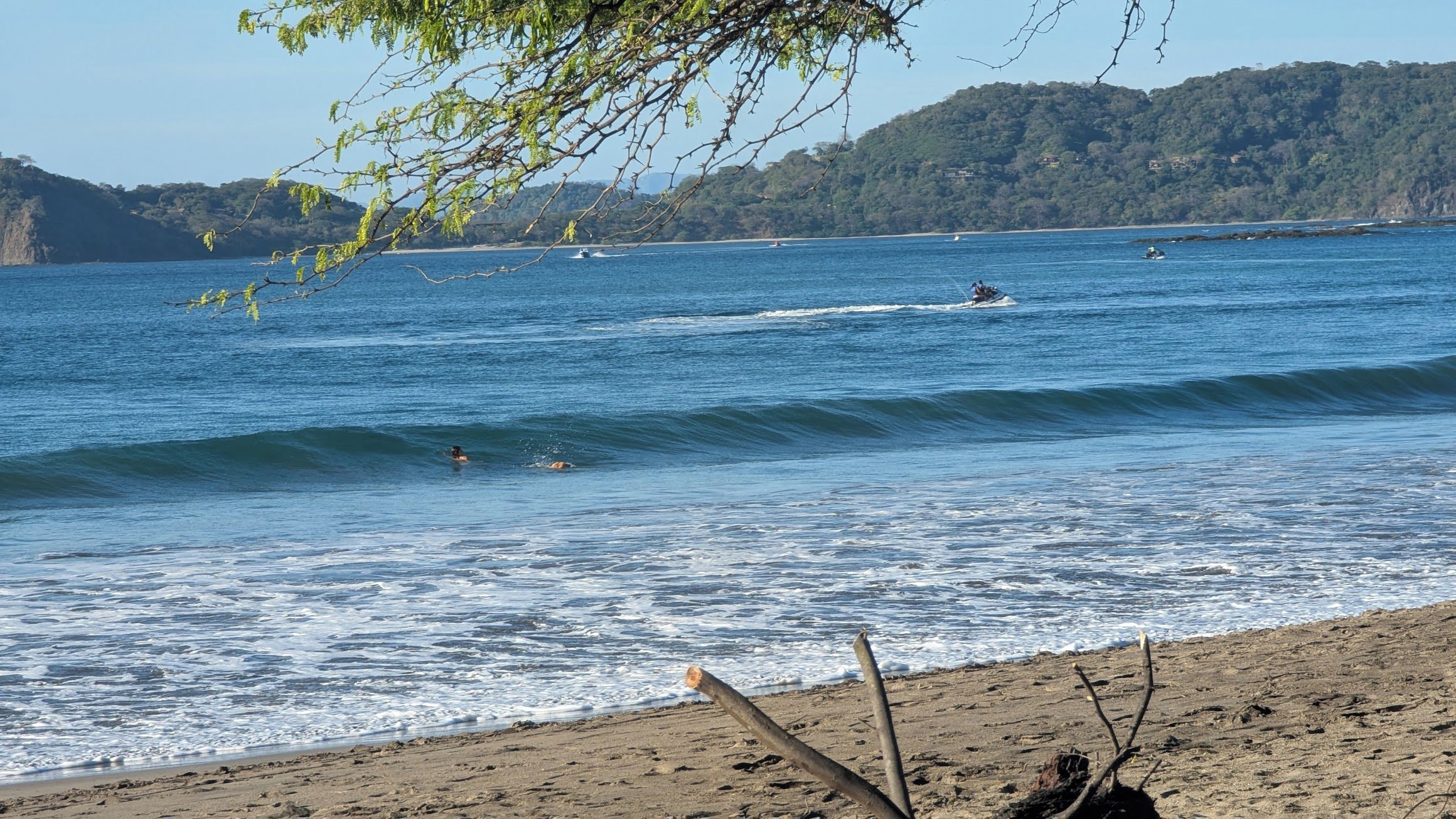 A beach with waves, a boat, and a forested hill in the background.
