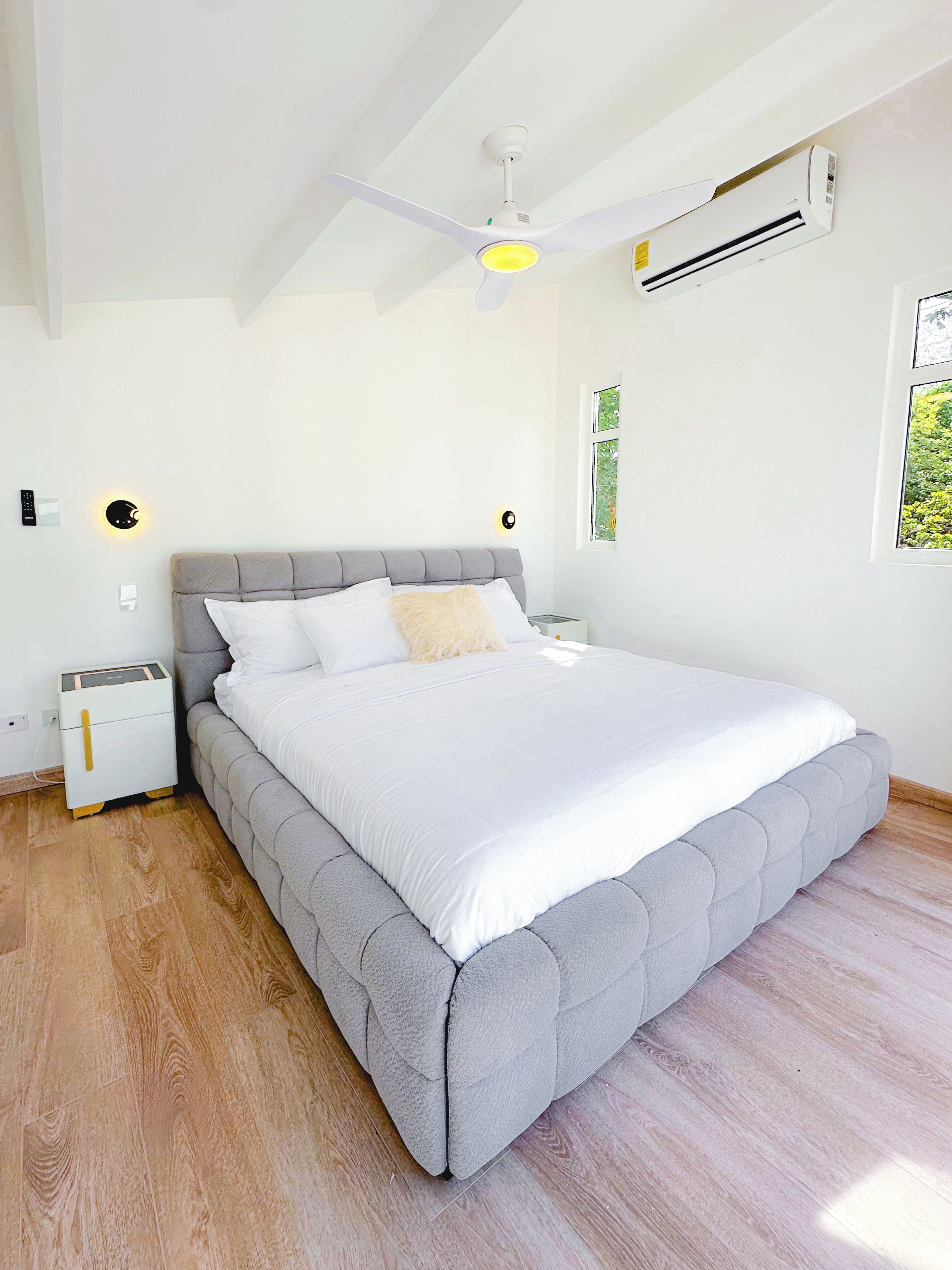A bedroom with a large bed, a ceiling fan, an air conditioner, a nightstand, and a bedside table.