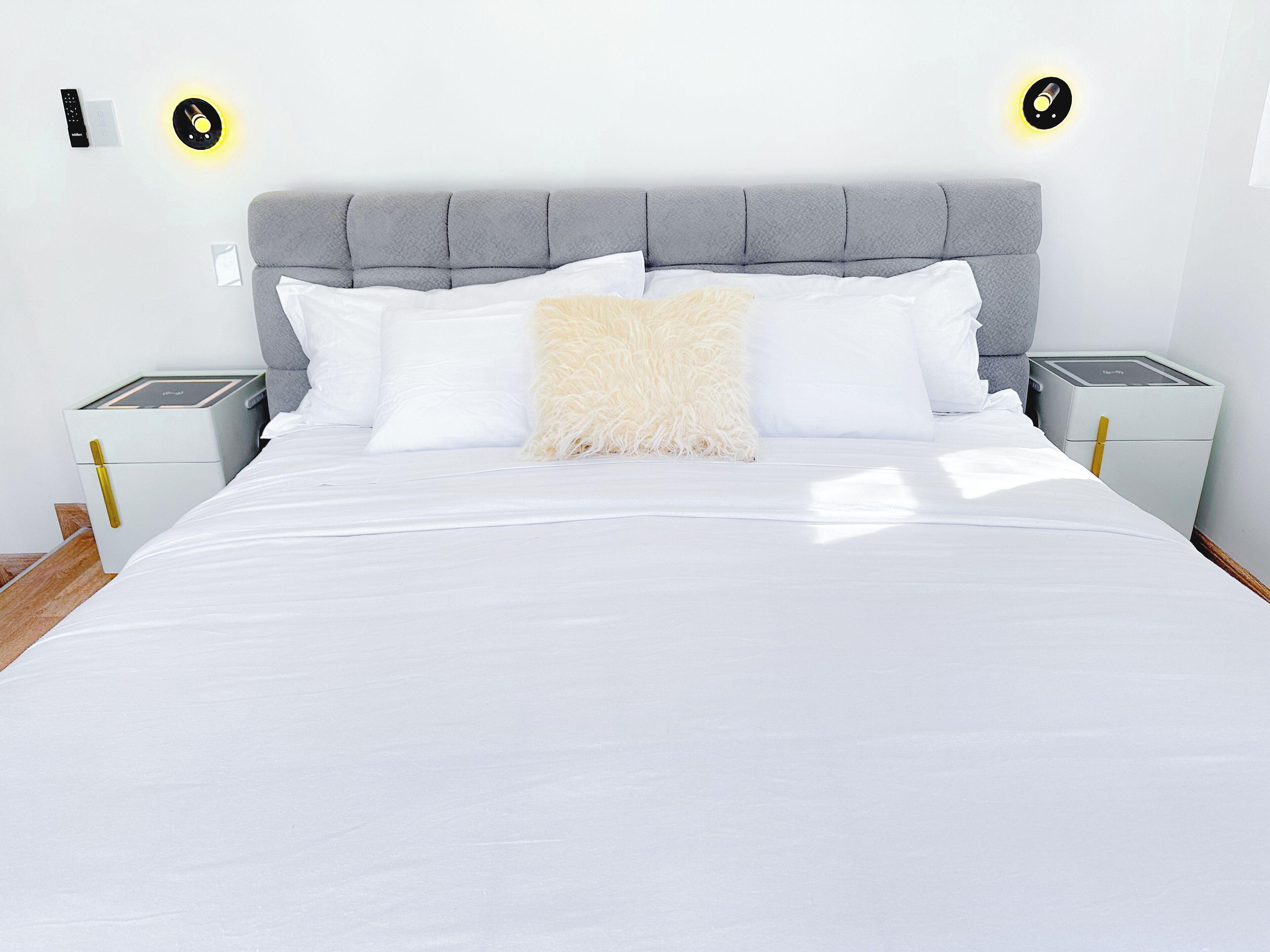 A neatly made bed with white linens, a fluffy beige pillow, and a grey upholstered headboard. There are two bedside tables with lamps, and the room has a clean, minimalist design.