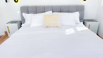 A neatly made bed with white linens, a fluffy beige pillow, and a grey upholstered headboard. There are two bedside tables with lamps, and the room has a clean, minimalist design.