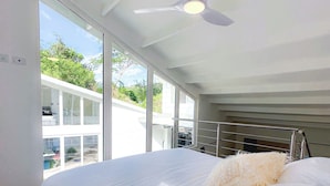 A modern bedroom with a large bed, a ceiling fan, and large windows offering a view of the outdoors.