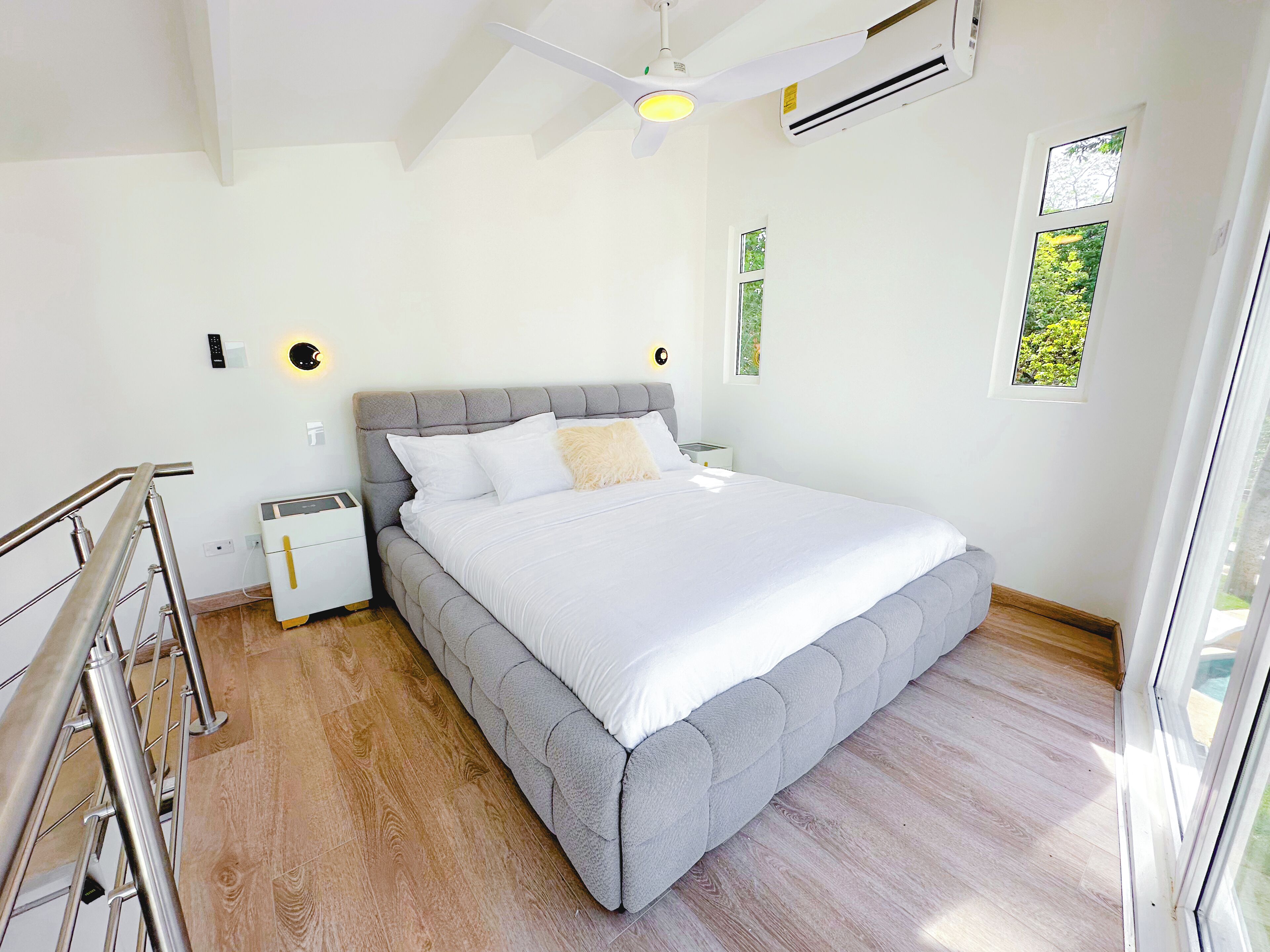 A modern bedroom with a large bed, a nightstand, a ceiling fan, and a window with a view of greenery.