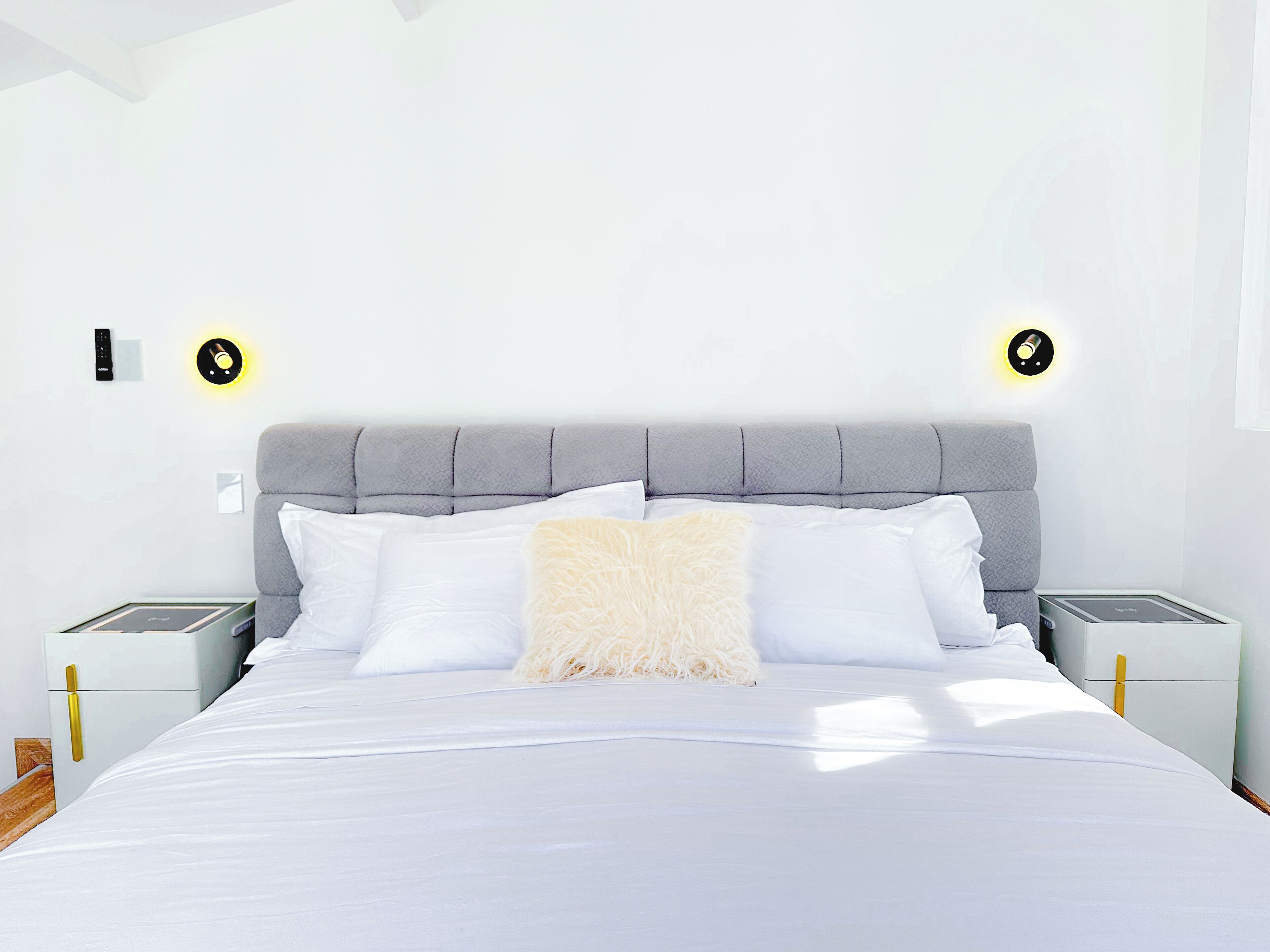 A modern bedroom with a grey upholstered headboard, white bedding, and two bedside tables with built-in lamps.