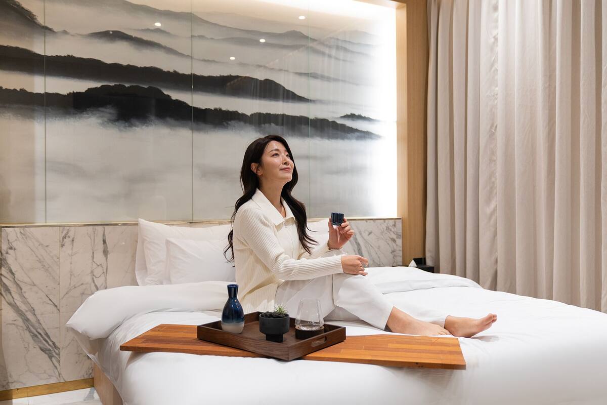 A woman sitting on a bed with a tray of items, a painting of mountains on the wall, and a curtain to the right.