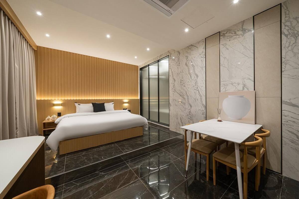 A modern hotel room with a bed, bedside table, and a desk with chairs.