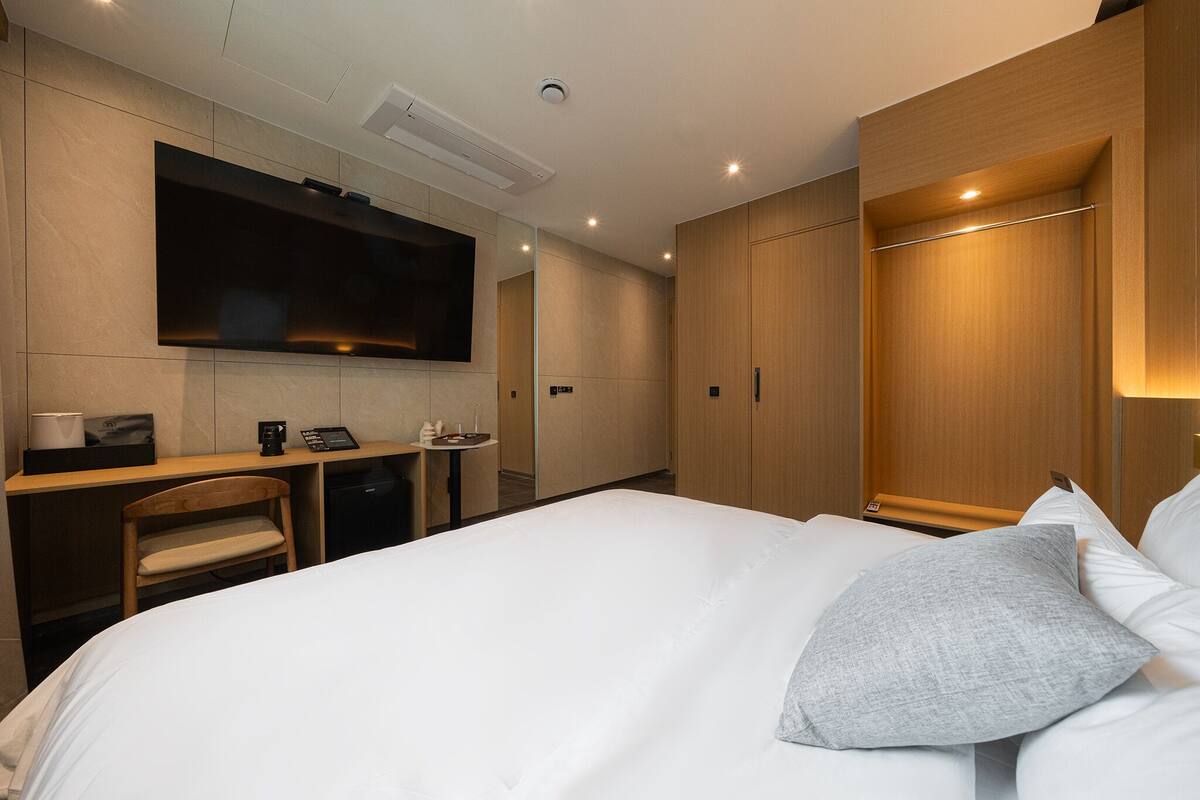 A modern hotel room with a large bed, a desk with a chair, a flat-screen TV mounted on the wall, and wooden wardrobes.