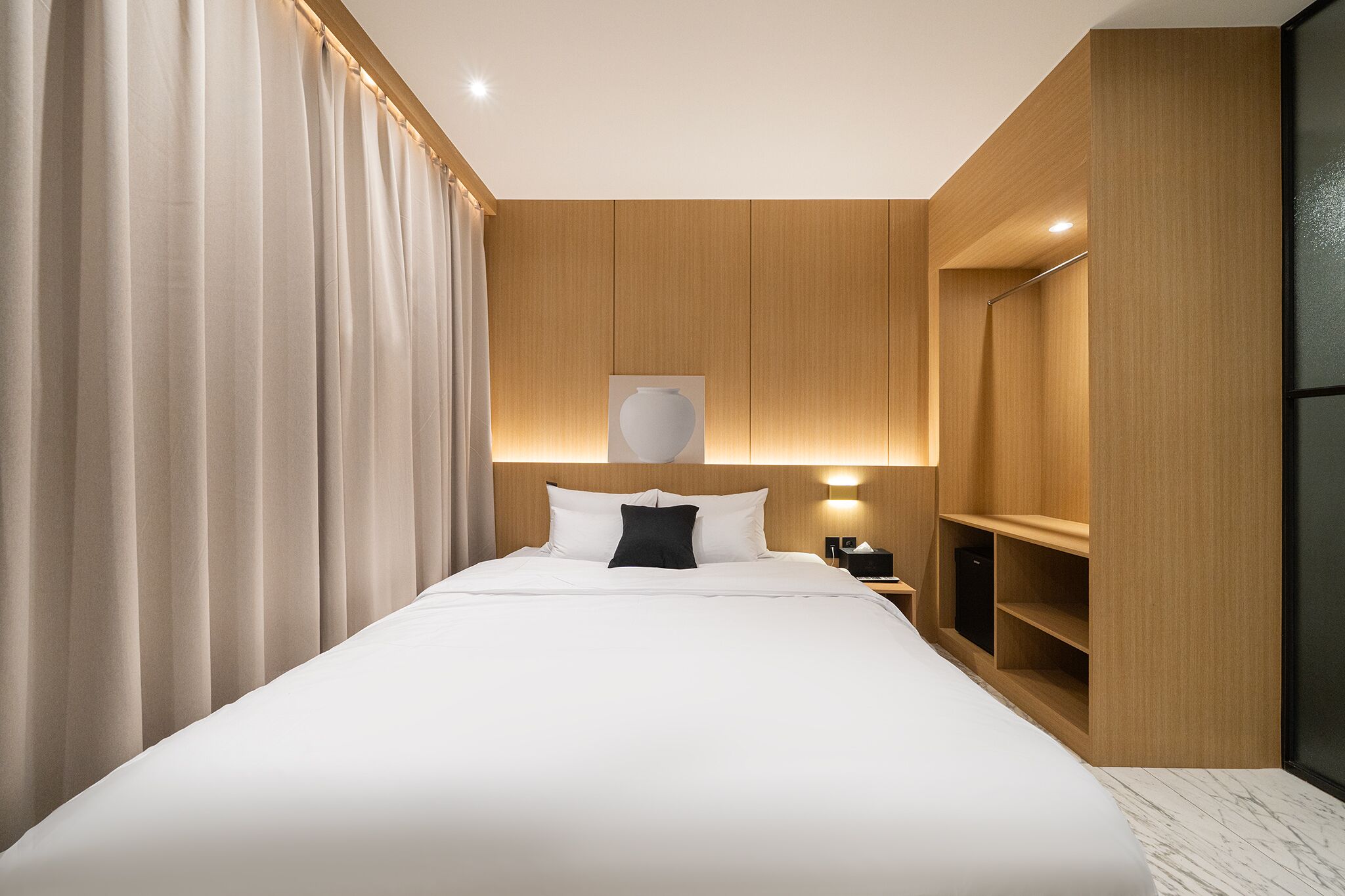 A modern hotel room with a large bed, wooden panel walls, a nightstand, and a built-in wardrobe.