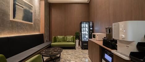 A modern lounge area with a green sofa, a black coffee machine, a microwave, and a digital coffee dispenser.