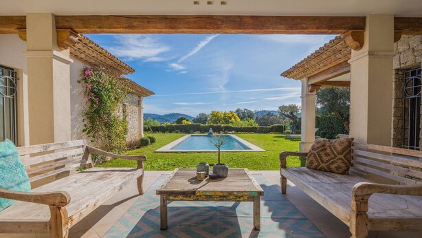 Un área de estar al aire libre con dos bancos de madera, una mesa y vistas a una piscina y vegetación.