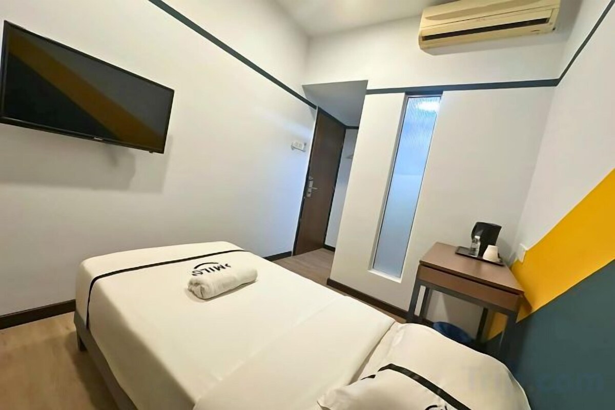 A hotel room with a bed, a flat-screen TV mounted on the wall, a small table with a coffee maker, and a window providing natural light.