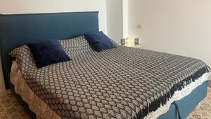 A neatly made bed with a patterned bedspread and two pillows, a bedside table with a lamp, and a wall-mounted light switch. - Cortina 2-24 Comfortable two-room apartment facing the sea (Comacchio)