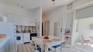 A compact kitchen with a washing machine, a dining table with chairs, and a patterned floor. - Cortina 2-24 Comfortable two-room apartment facing the sea (Comacchio)