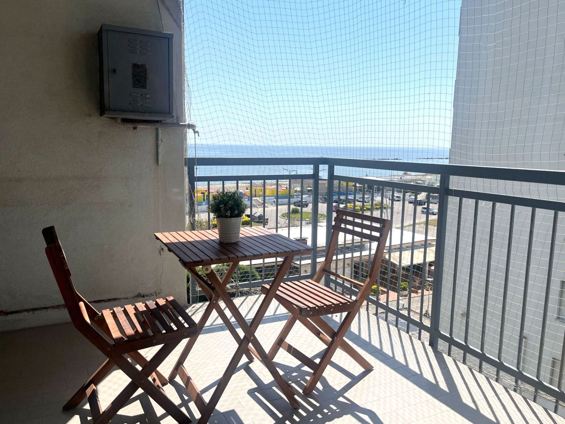 A balcony with a wooden table and two chairs, a potted plant, and a view of the sea and cityscape.