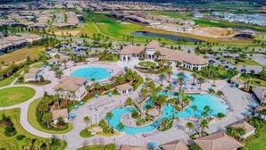 An aerial view of a residential area with two large swimming pools, a golf course, and a central pavilion.