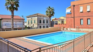 A rooftop swimming pool with a clear blue pool, surrounded by a metal railing. There are palm trees and a mix of residential buildings in the background. - TWO-ROOM APARTMENT SLEEPS 5 IN THE CENTRAL AREA (San Vincenzo)