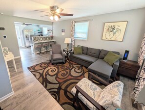A living room with a sofa, chairs, a coffee table, a rug, a ceiling fan, and a kitchen area in the background.