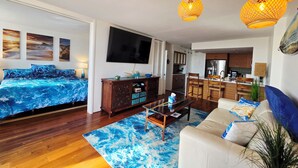 A modern living room with a sofa, coffee table, and a TV. There is a kitchen area visible through an open door. - Ocean and Marina View Suite, Spacious, Quiet and Peaceful with Amazing Sunsets (Honolulu)