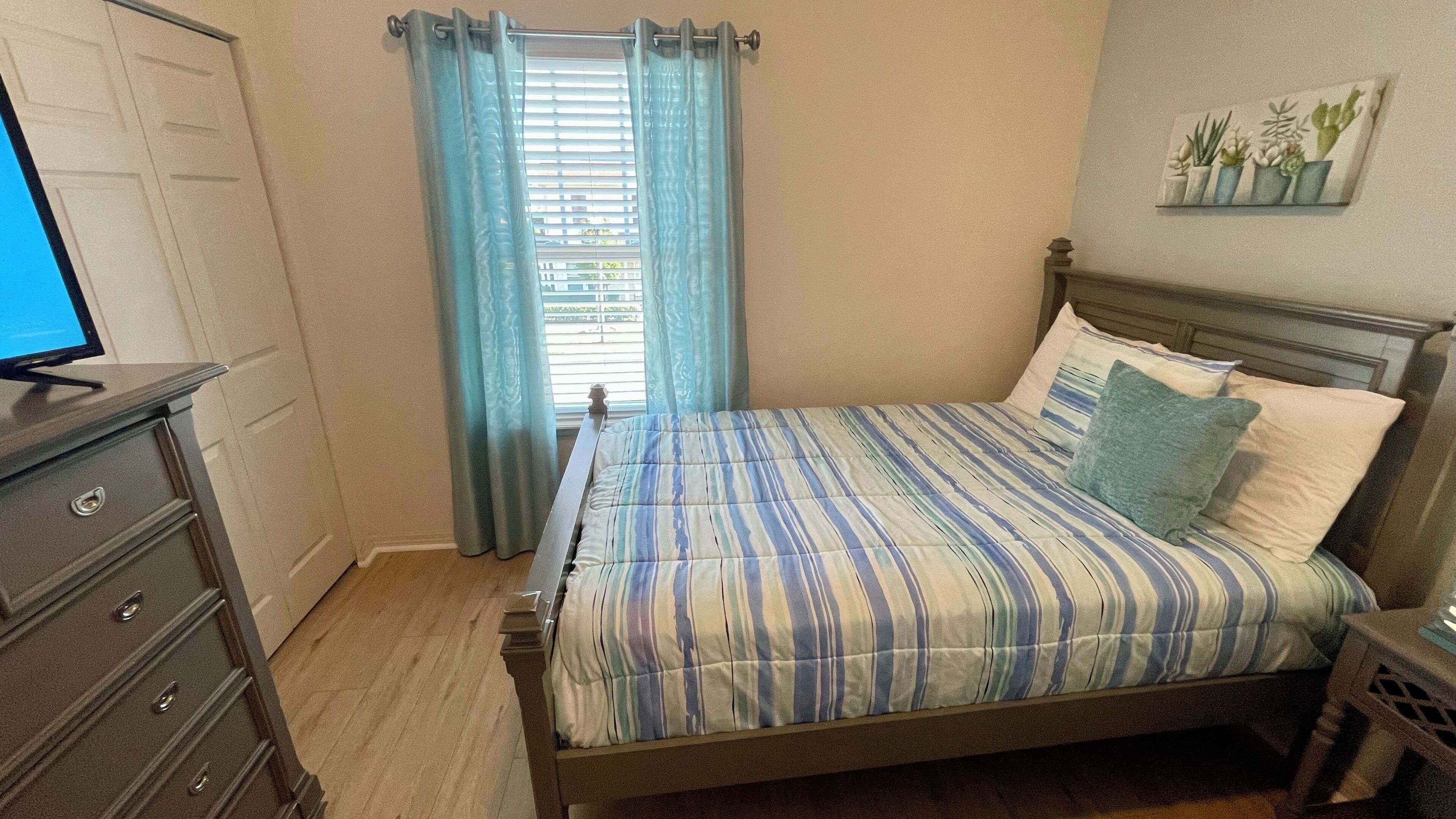 A bedroom with a bed, a dresser, a television, and a window with curtains.