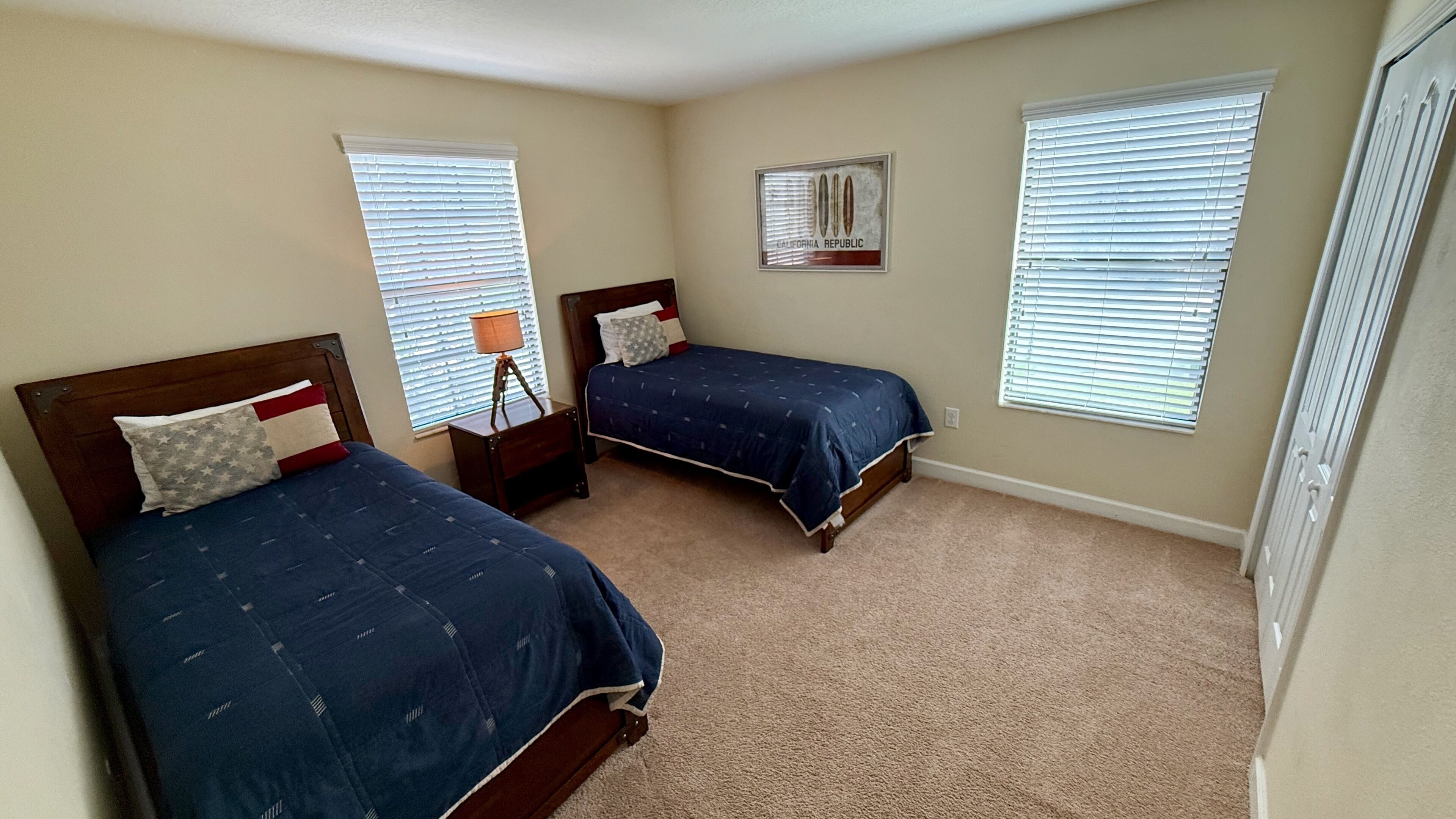 A bedroom with two beds, each with a headboard, a nightstand with a lamp, and a window with blinds.