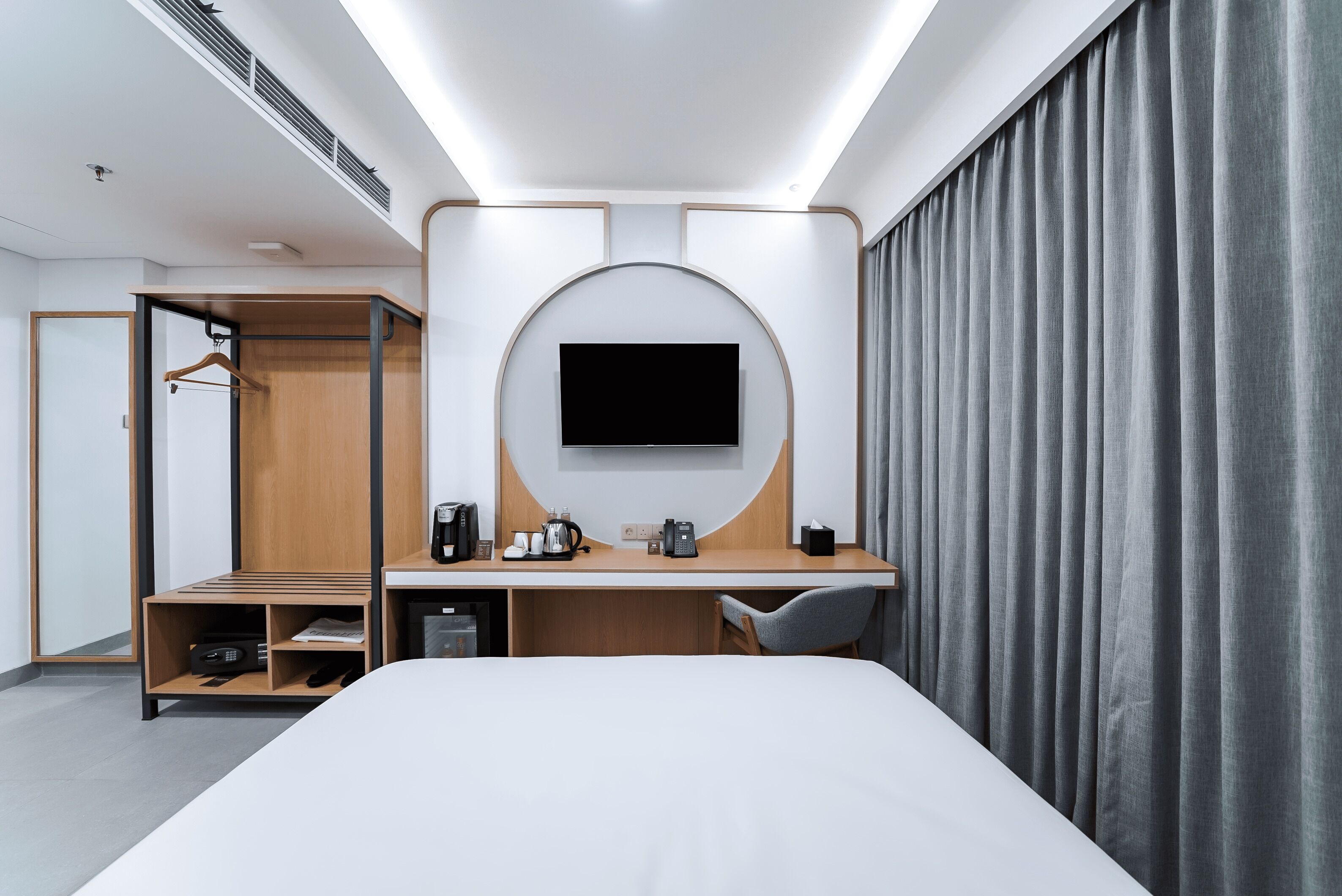 A modern hotel room with a bed, a desk with a flat-screen TV, a wardrobe, and a large window with curtains.