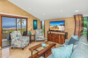 A living room with a large TV, a sofa, a chair, a coffee table, and a balcony with a view of the ocean.