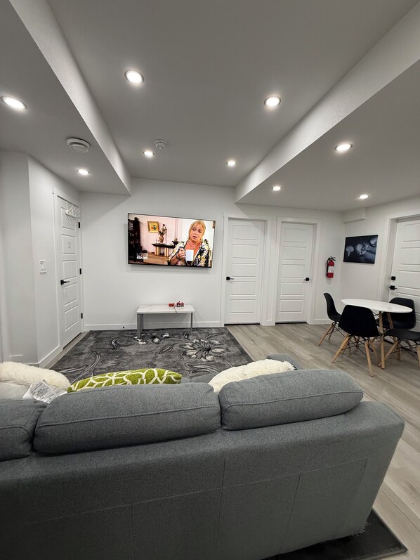 A modern living room with a grey sofa, a flat-screen TV mounted on the wall, a dining area with a table and chairs, and a patterned rug on the floor.