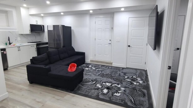 A modern living room with a black sectional sofa, a flat-screen TV mounted on the wall, and a kitchen area with white cabinets and a microwave. - Carrington Cozy Suite, Calgary (Calgary)