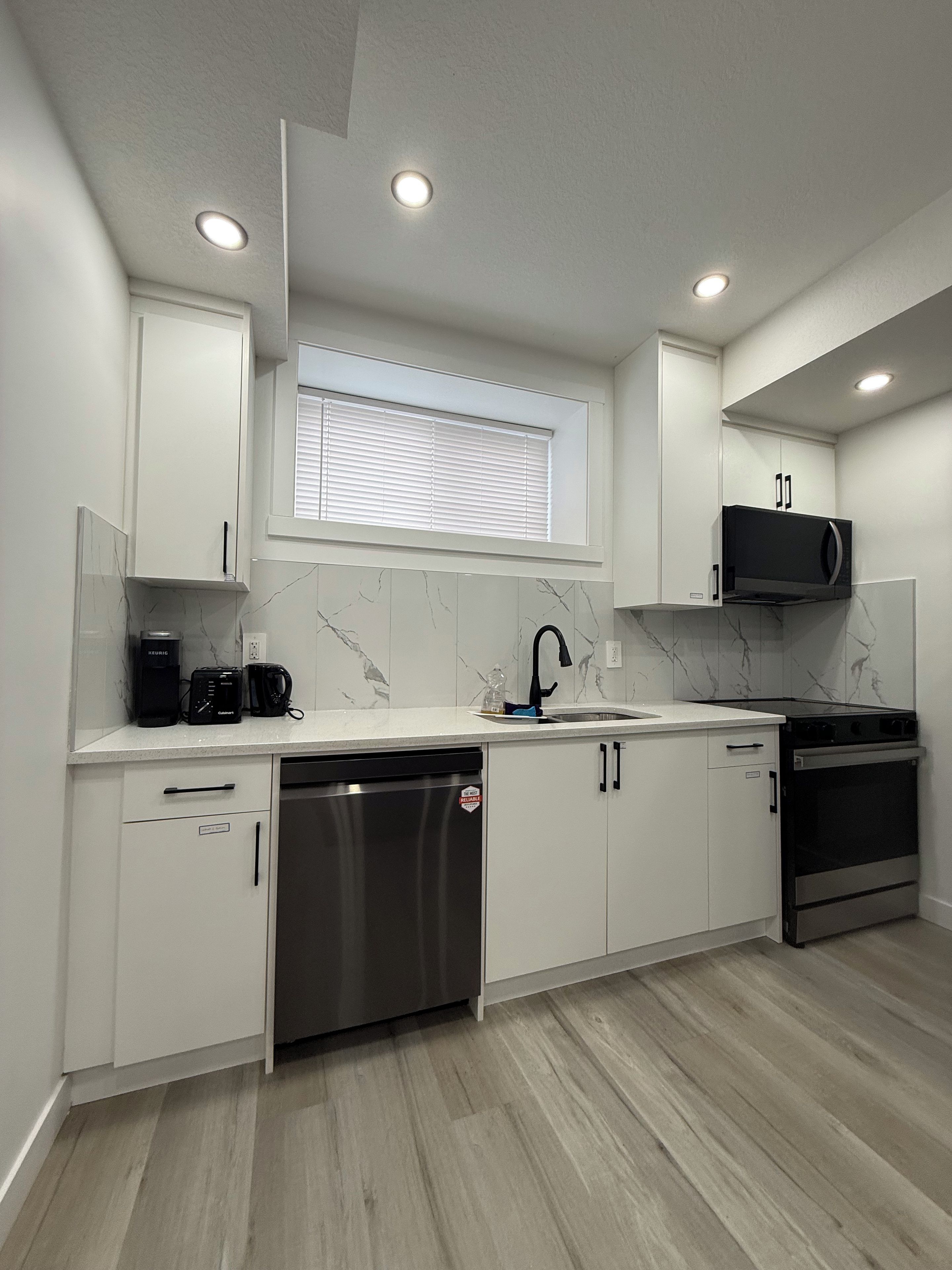 A modern kitchen with white cabinets, a built-in microwave, a stainless steel dishwasher, and a window with blinds.