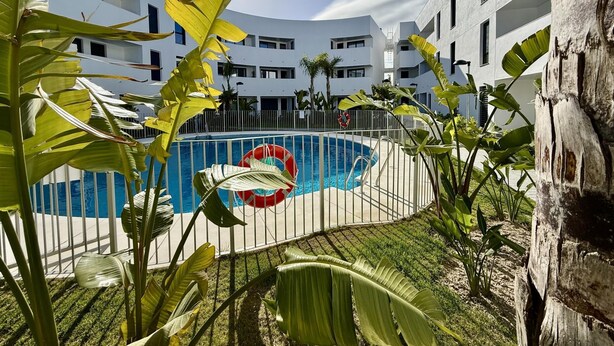 Une piscine entourée d’un bâtiment blanc doté de balcons, lui-même entouré de plantes tropicales.