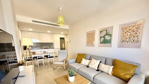 Un salon moderne avec un canapé gris, une table basse en bois et un coin repas comprenant une table et des chaises blanches. Des œuvres d’art encadrées ornent le mur, et une suspension jaune éclaire l’espace.