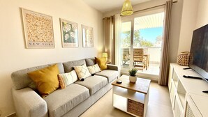 Un salon moderne avec un canapé gris, une table basse et un téléviseur posé sur un meuble blanc. Trois reproductions botaniques encadrées ornent le mur, et on aperçoit un balcon avec des meubles d’extérieur à travers la porte vitrée.