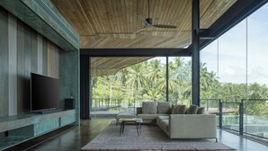 A spacious living room with a large flat-screen TV, a sectional sofa, and a view of palm trees through large windows. - Kelapa House (Koh Samui)