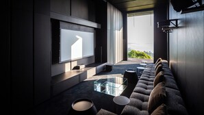 A modern living room with a large flat-screen TV mounted on the wall, a glass coffee table, a sofa with cushions, and a view of the outdoors through a large window. - Kelapa House (Koh Samui)