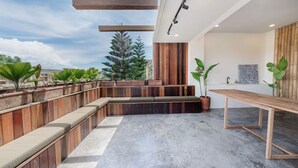 A modern outdoor seating area with wooden benches and planters, a concrete floor, and a view of the cityscape. - Modern 2BR Villa Rooftop & Pool (Kuta Utara)