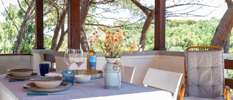 A covered outdoor dining area with a table set for two, a wicker chair, and a decorative vase with flowers.