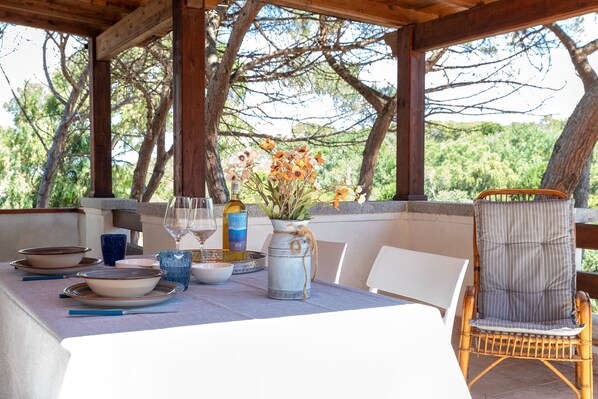 A covered outdoor dining area with a table set for two, a wicker chair, and a decorative vase with flowers.