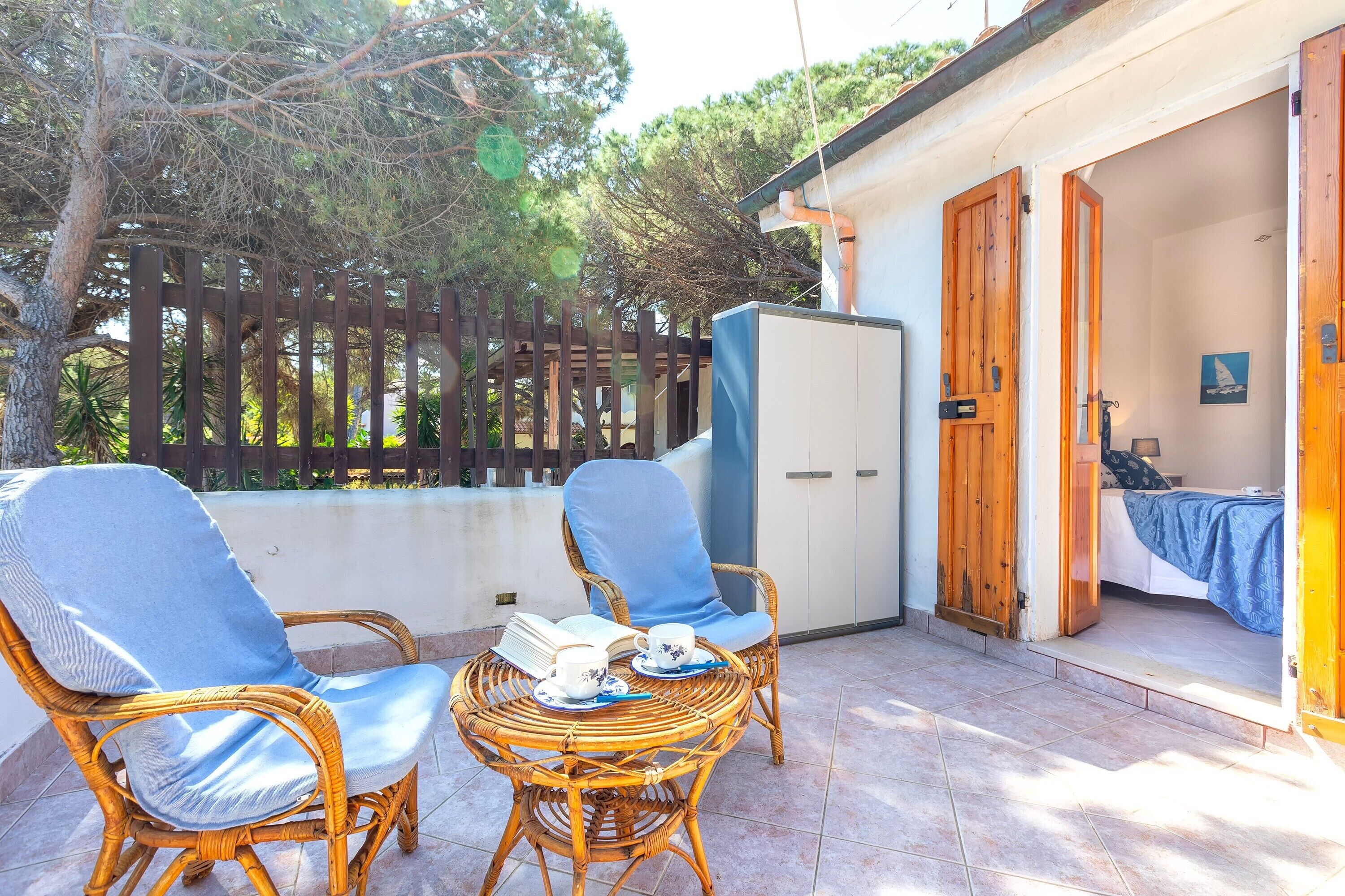Un balcon avec des fauteuils en osier, une table avec un livre et une tasse, et un lit recouvert d’une couverture bleue dans la pièce adjacente.